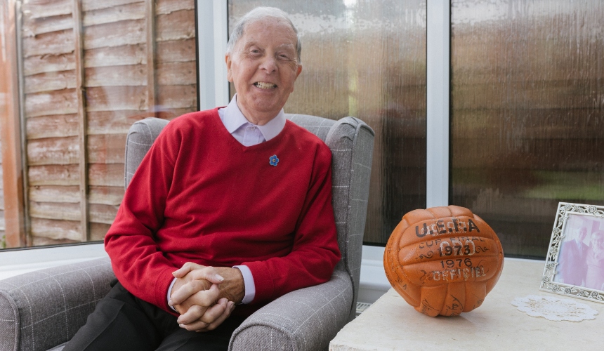 Bob sits on a couch smiling beside his football signed by the 1970s Liverpool football players