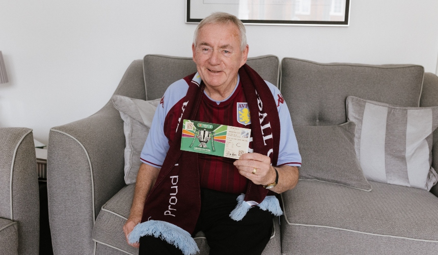 Peter sits on a couch wearing his Aston Villa Scarf, holding up a ticket