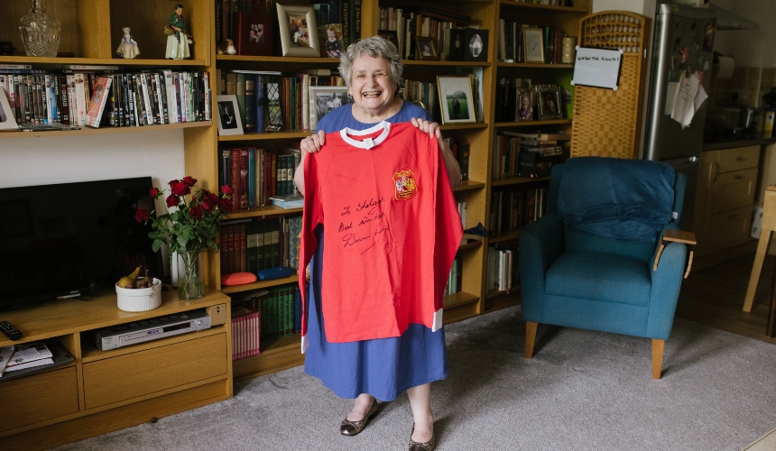 Shelagh stands in her living room, smiling and holding up a shirt gifted to her by Denis Law