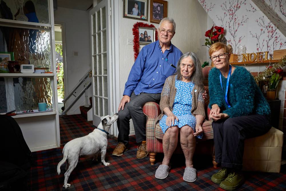 A woman with dementia sits on a sofa chair beside her husband, who is supporting her. A dementia advisor sits next to them.