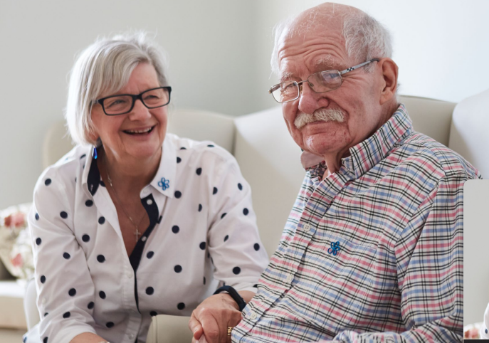 A man and a woman sit together on a sofa, smiling and holding hands.