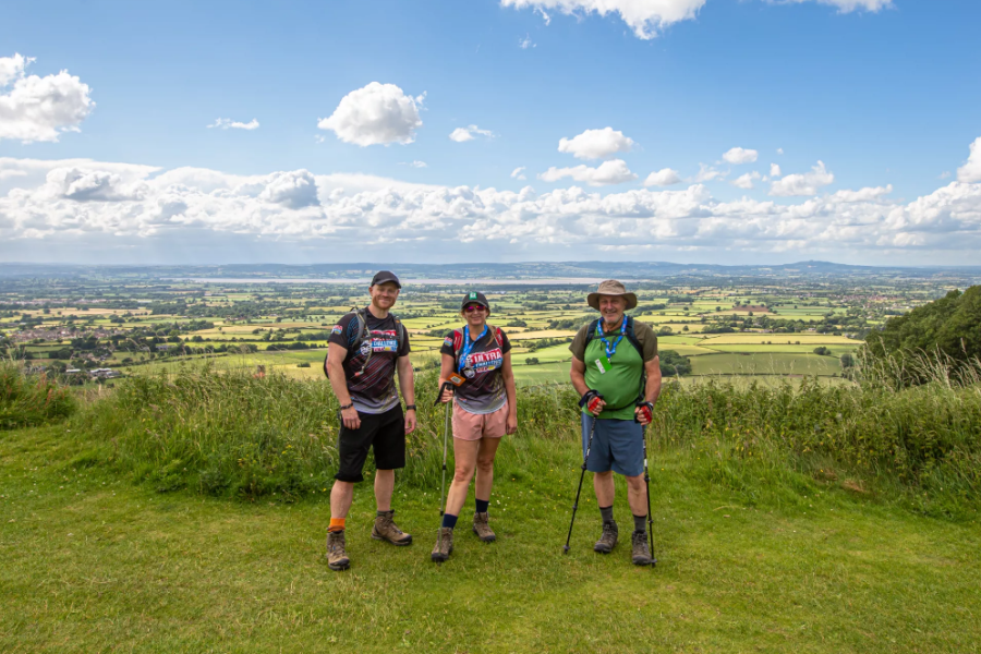 Three trekkers stand atop a hill with a country vista in the background