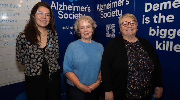 Three women smiling at the camera stood in front of Alzheimer's Society branding.