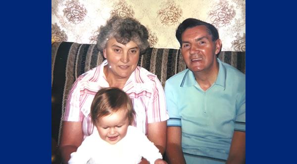 Emma as a baby being held by her Nan and Granddad sat on a brown stripy sofa