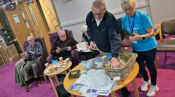 Man reaches into a hamper on a table, he's stood next to a woman in an Alzheimer's Society t-shirt, behind are three people sat in purple chairs.