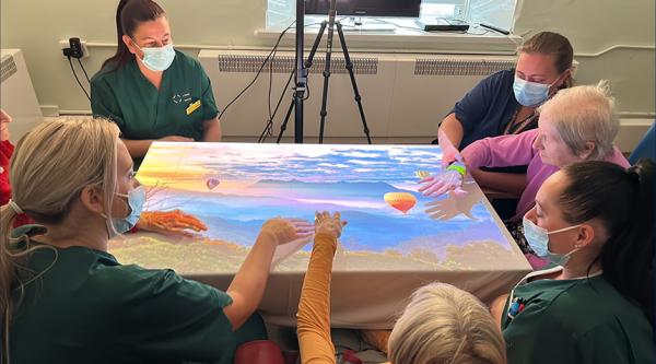 Carers and people with dementia sat around a table looking and pointing at light game projected onto the tabletop