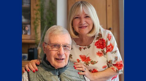 Maria is stood up and wearing a flowery top, with her hands placed on Roy who is sat down and wearing a green gilet