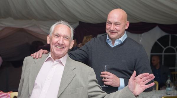 An older man waves at the camera, his middle-age son stands behind him with a hand on his shoulder.