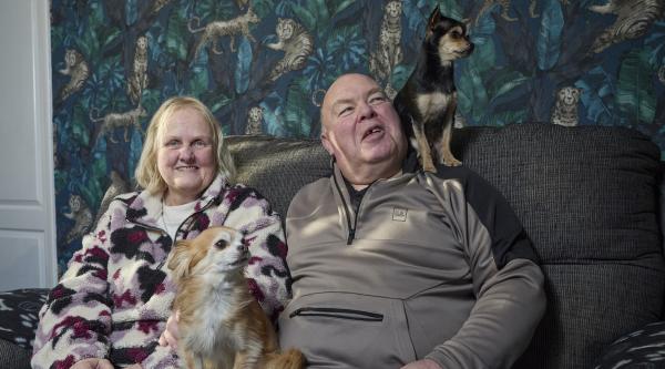 A man and woman sit on a grey sofa. There is a small brown and white dog sat between them, and small black and brown dog sits on the man's shoulder