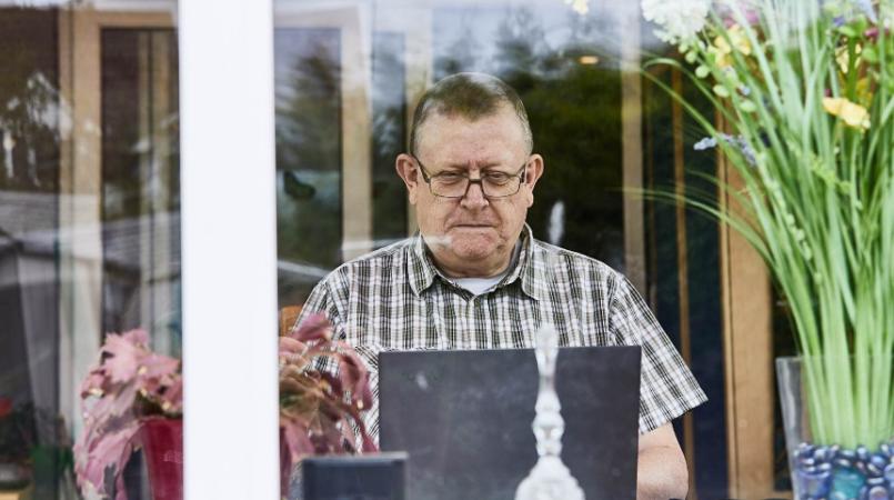 An older man sits at the window in his living room, reading something on his laptop