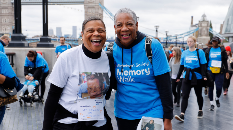 2 women smiling with Tower Bridge in the background, one in a blue Memory Walk t-shirt and one in a plain t-shirt with a picture of a loved one.