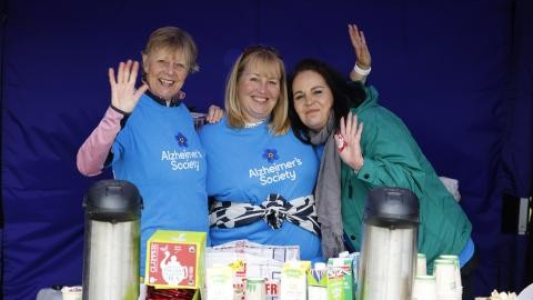 Three women at an outdoor hot drink counter wave towards the camera