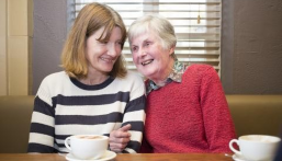 An older and younger woman sit arm in arm enjoying their coffee.