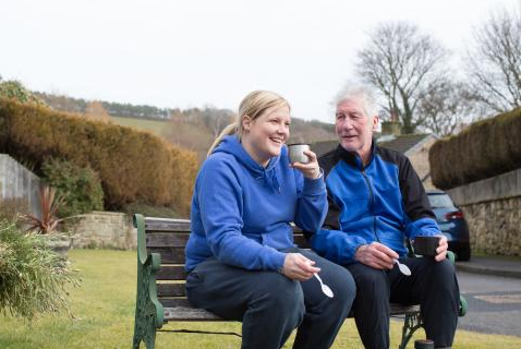 A man and a woman in blue Alzheimer's Society hoodies sit on a park bench.