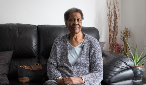 A woman sits patiently on her sofa, looking calmly to camera.