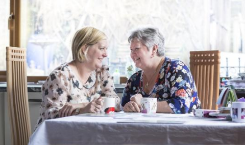 Two women sit closely together at a kitchen table with mugs of tea