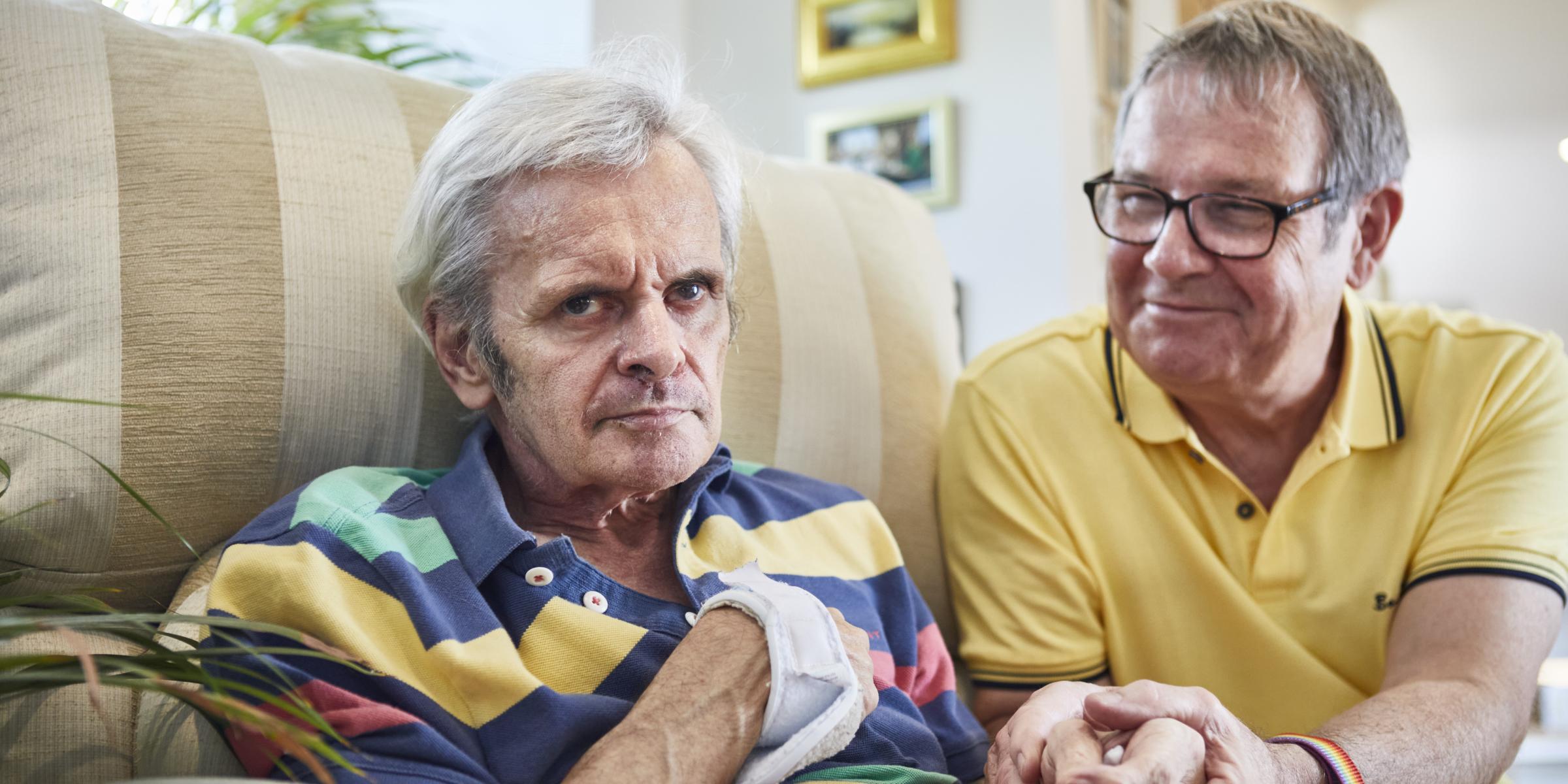 A man in a yellow top holds the hand of a man sat in a beige sofa wearing a colourful stripy top